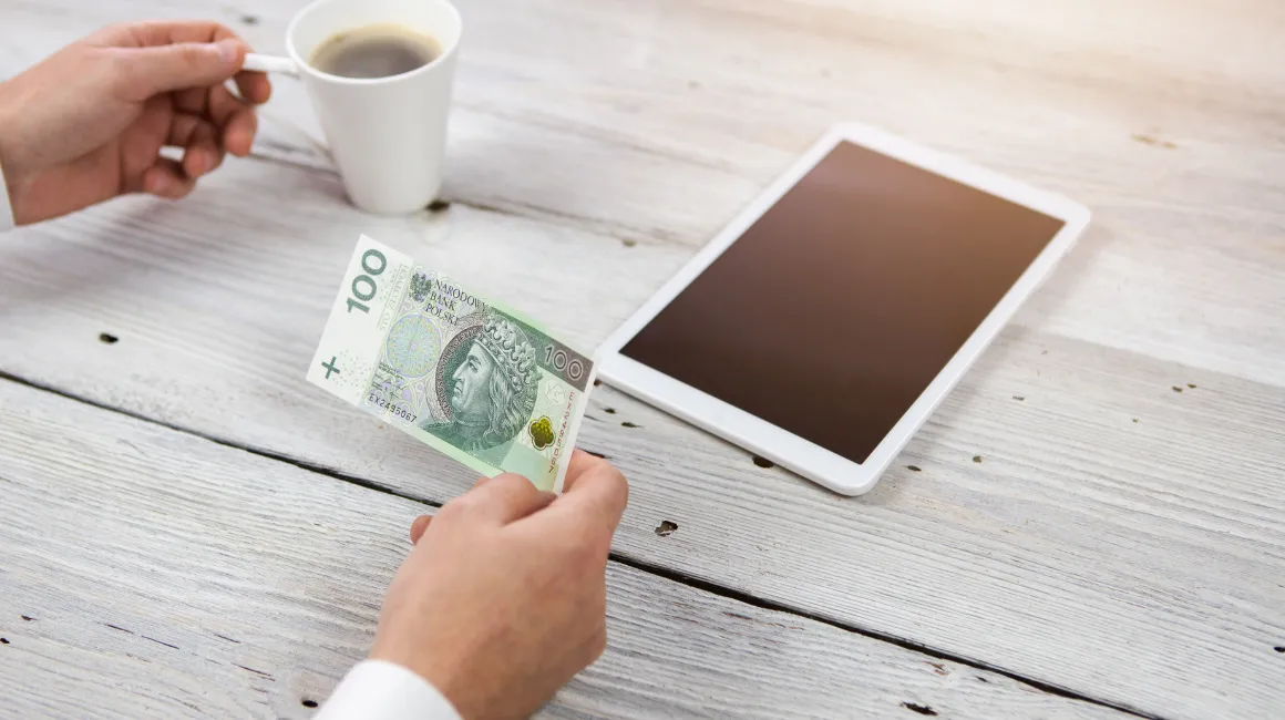 Osoba trzymająca banknot 100 złotych przy filiżance kawy i tablecie